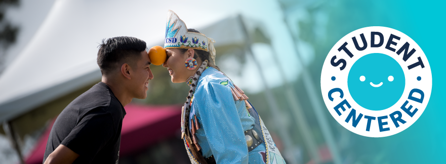 1 of 2, Two students holding an orange between their foreheads at the annual Powwow